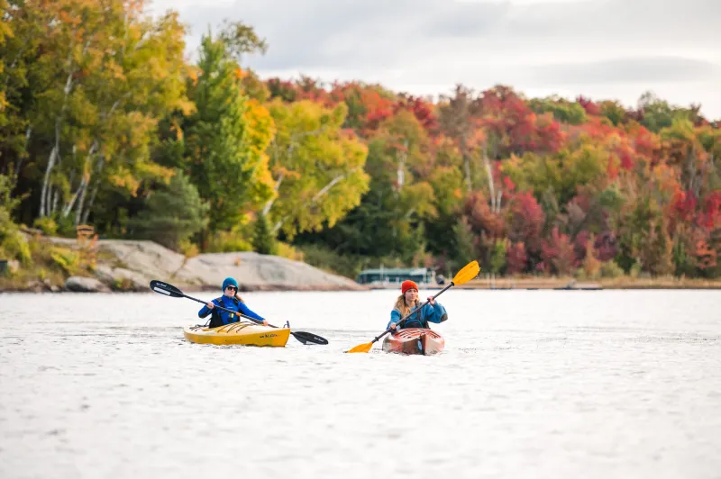Two women paddle kayakas along a lake with fall foliage along the shoreline