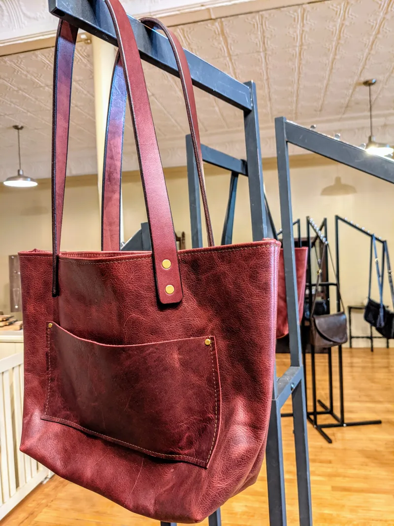 A handmade burgundy leather tote bag hangs on a rack in a shop.