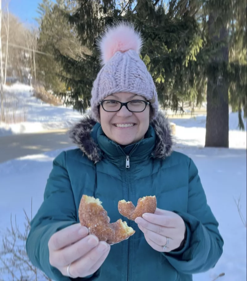 A woman in pink winter hat and green coat holds out half of a cinnamon sugar donut.