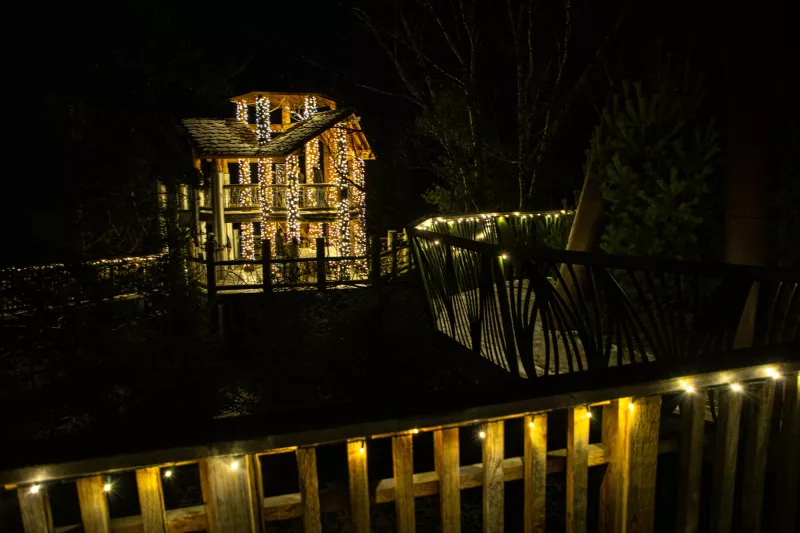 An elaborate wooden treehouse covered in white holiday lights at night.