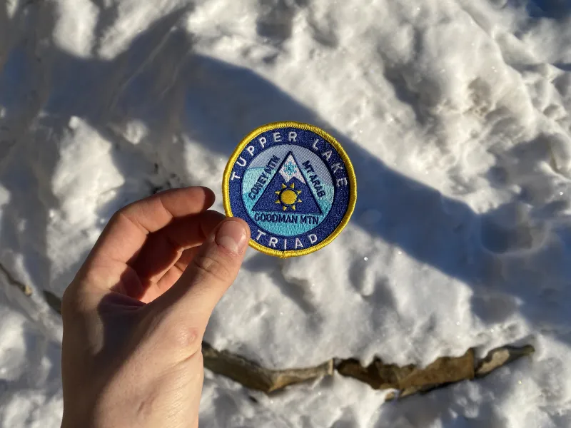 A hand holds out the Tupper Lake Triad challenge patch in front of the snow.