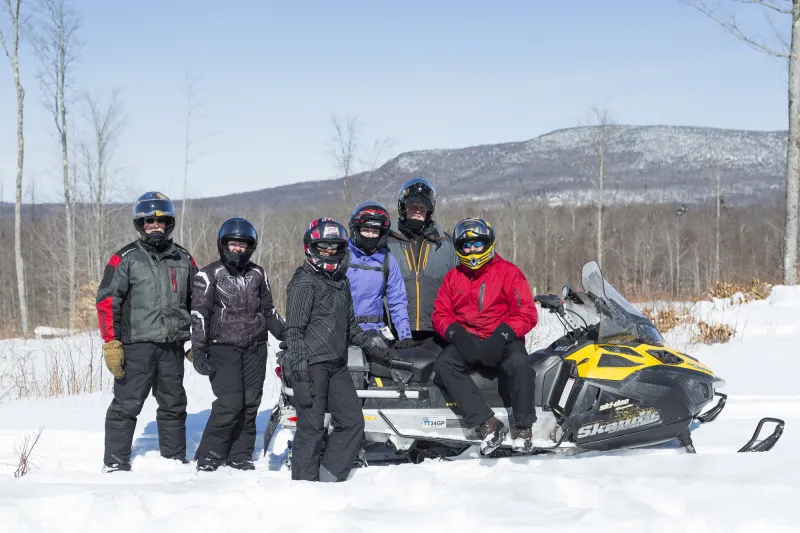 A group of six people pose with a snowmobile along a snowy trail.