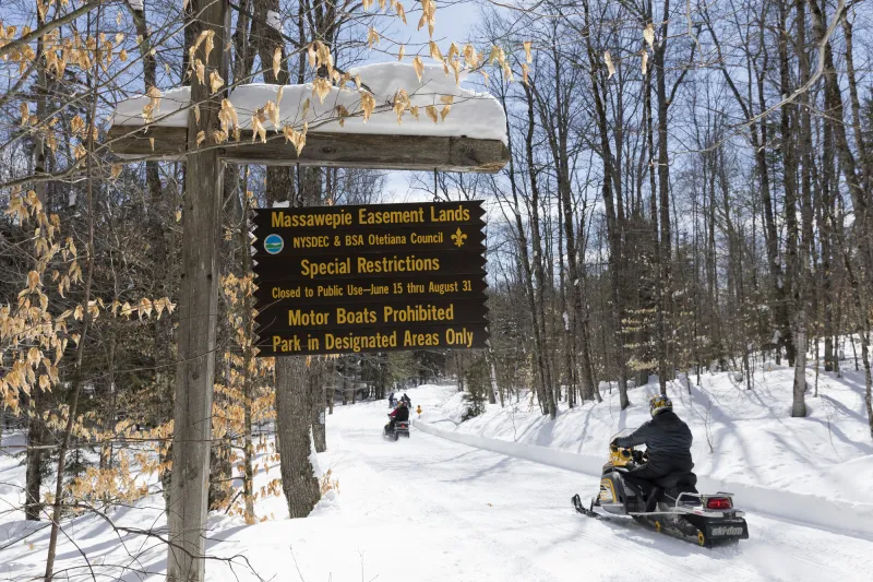A group of snowmobilers ride down a groomer trail near a DEC sign for Massawepie conservation easement lands.