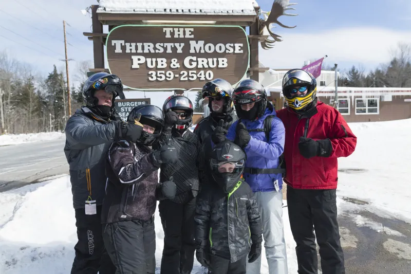 A group of snowmobilers smile outside a restaurant, with the sign in the background for The Thirsty Moose Pub.