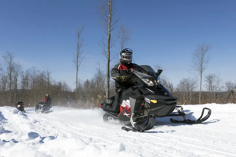 A snowmobiler in a red snowsuit on a black sled rides on a groomed trail in an open area with trees in the background.