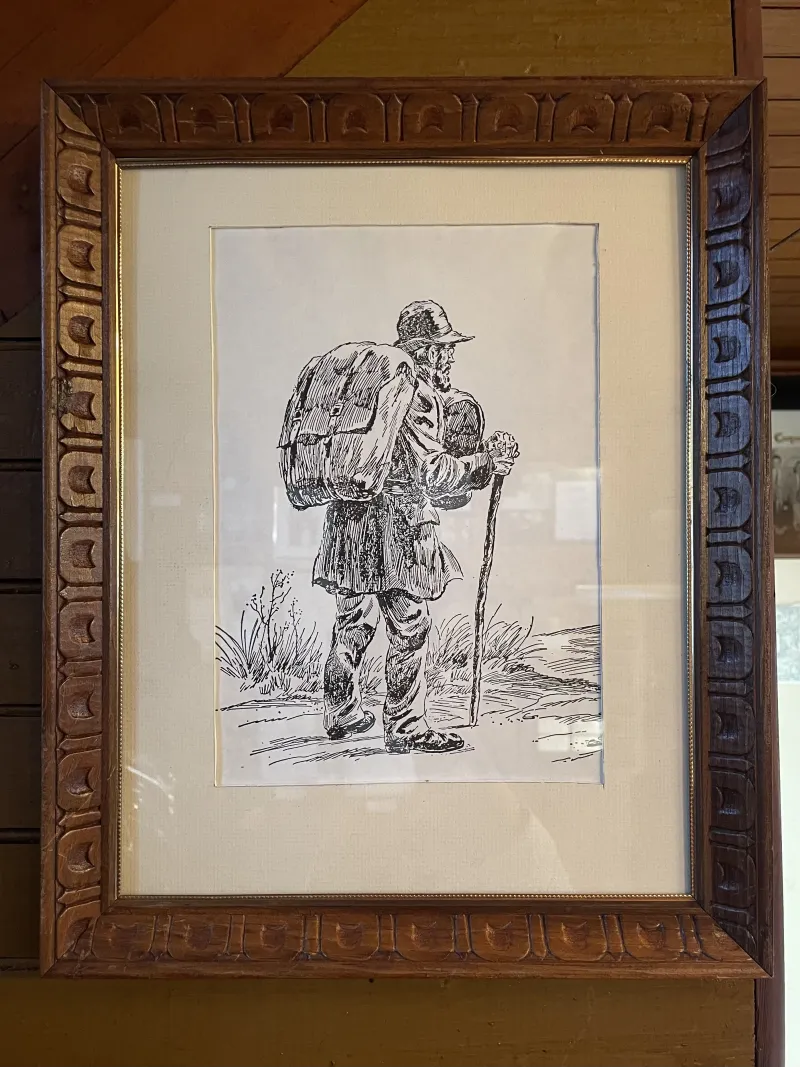 A framed black and white sketch of an old-fashioned peddler with walking stick and oversized pack.