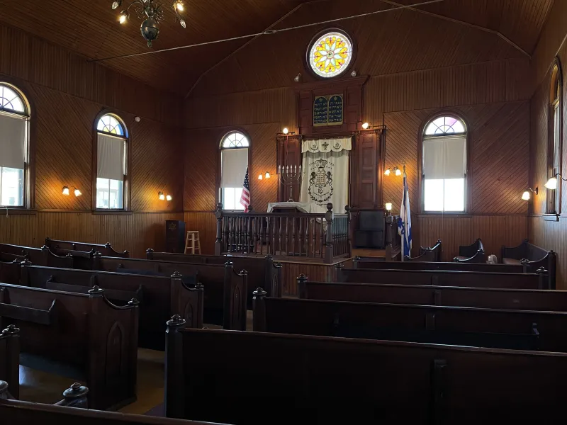 The interior of a early 20th century synagogue with wooden paneling and stained glass windows.