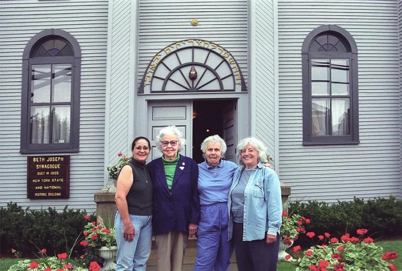 Four women stand side by side in front of an early 20th century synagogue.