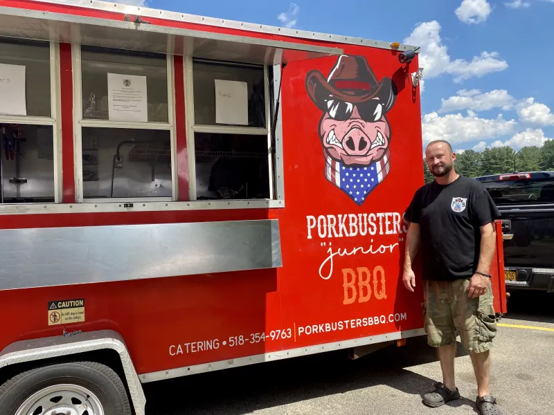 A man stands in front of his red food truck. A smiling pig image features on the truck.