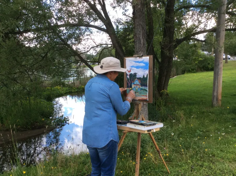 a woman paints a house scene on an easle by the lake.