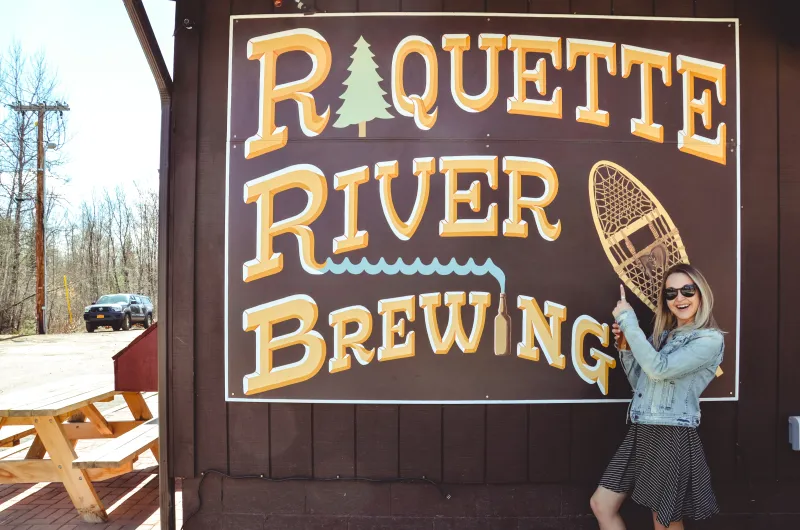 a woman poses with a giant sign that says "Raquette River Brewing."