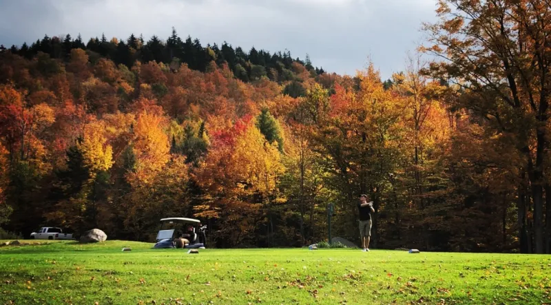 A golfer and cart in front of a forested hill covered in bright fall foliage.
