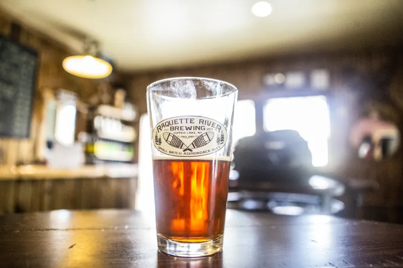 A glass of beer with the Raquette River Brewery logo etched on it sits on a bar.