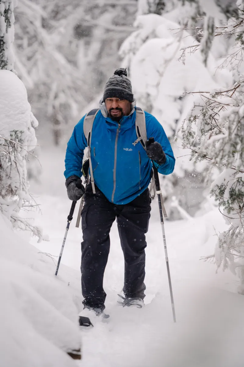 A man in a blue jacket snowshoes as it snows on a winter trail.