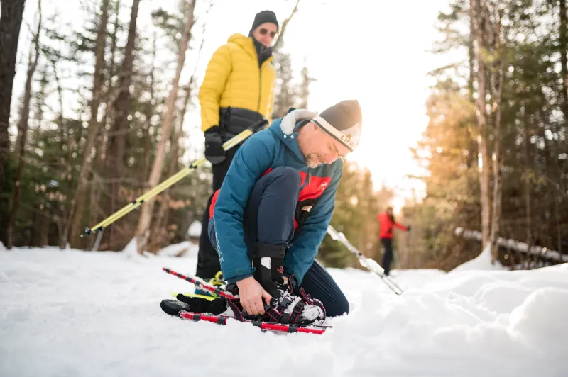 A man strap a snowshoe to his foot as his partner waits next to him