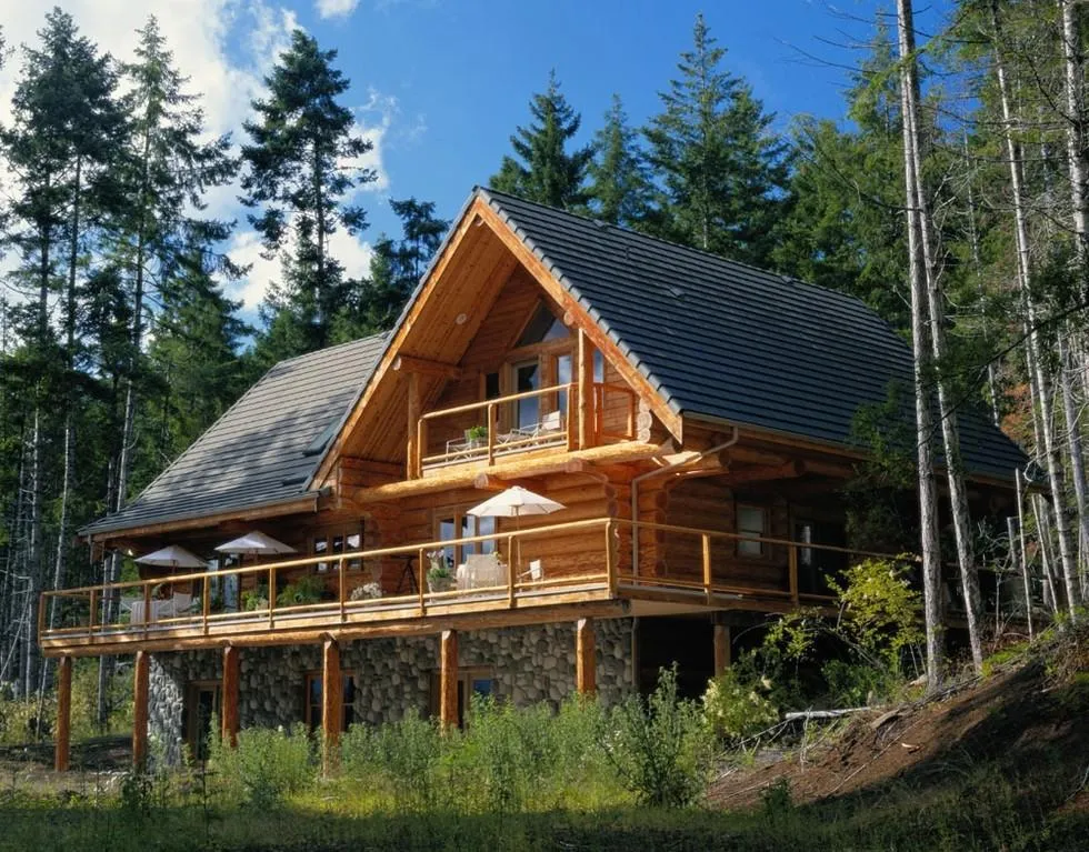 A newly-built log cabin vacation rental with dark roof, large open deck, and stone-walled basement sits in front of large pines