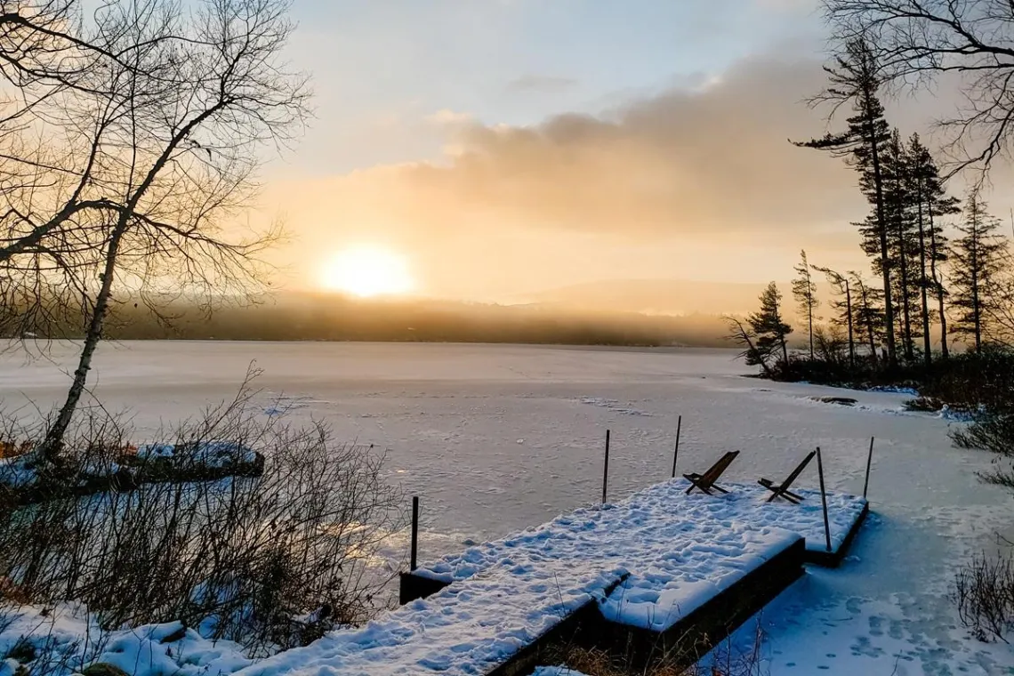 View from a vacation rental in Tupper Lake showcasing a snow-covered dock sitting on a frozen lake while the sun rises above the mountains in the background.