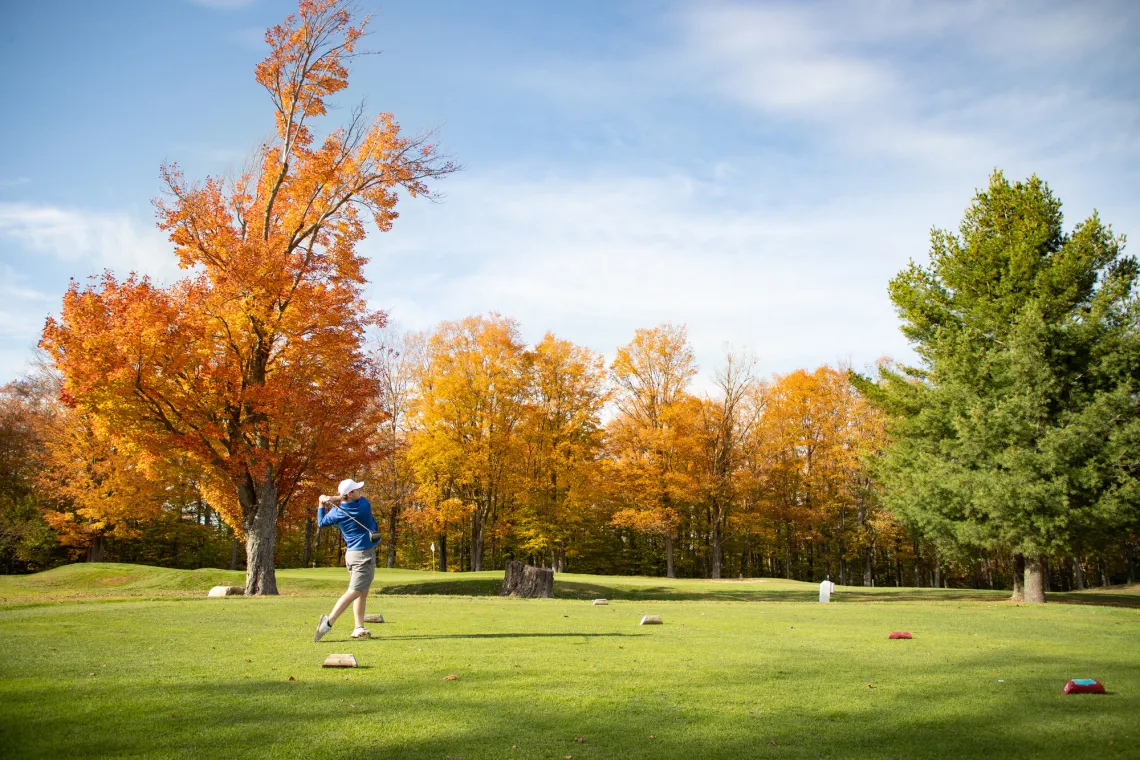 A golfer tees off in Tupper Lake surrounded by vibrant fall foliage in shades of red and orange.