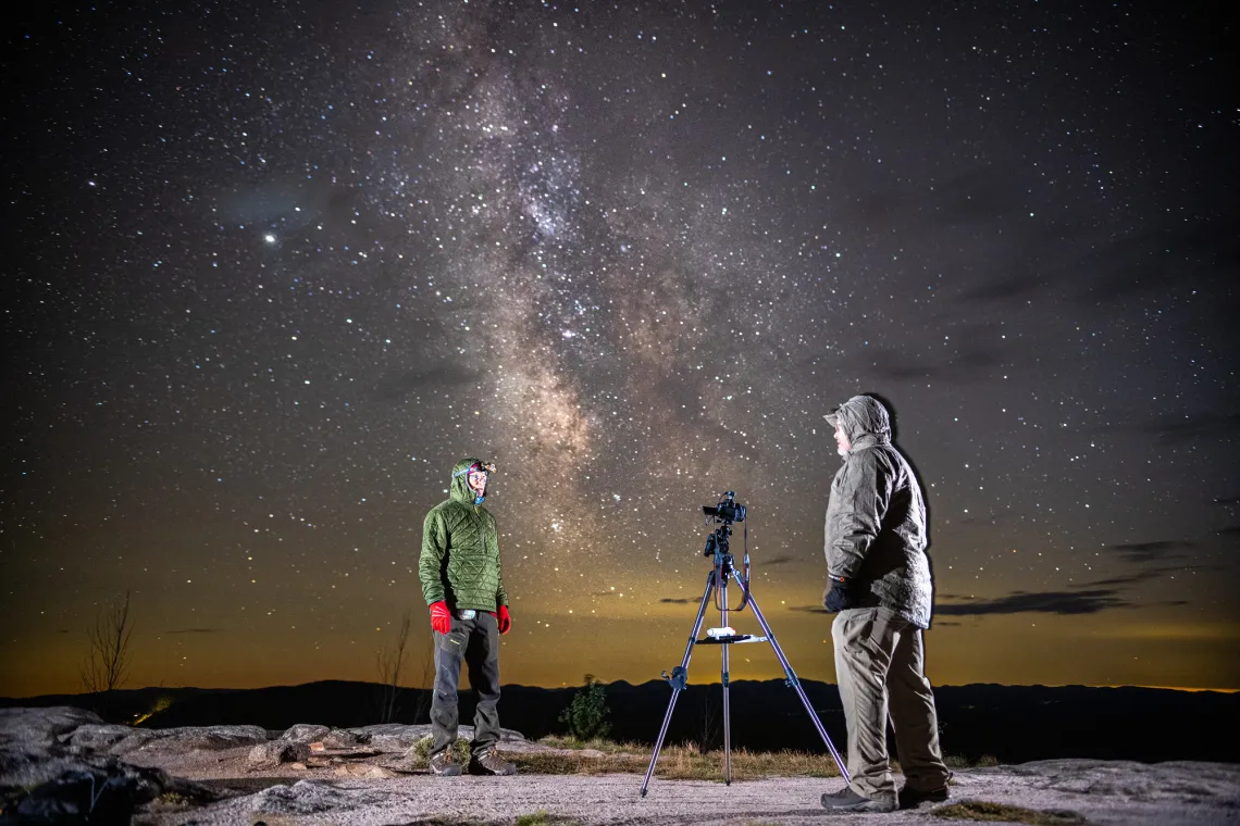 Two men stand on a rocky summit with the night sky and MIlky Way filling the sky behind them.