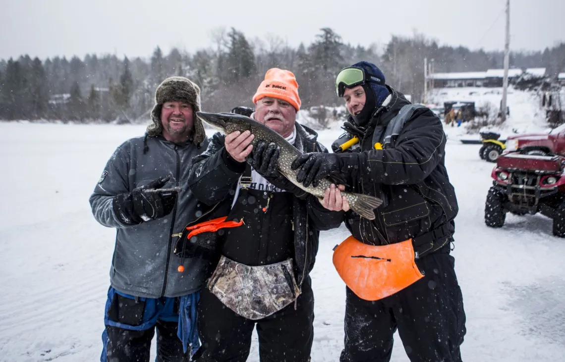 Three men on a frozen lake pose with their latest catch while snow falls around them