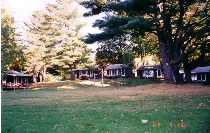 A brown house with white trim tucked in the pine forrest beyond a large lawn.
