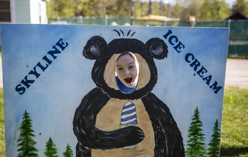 A child smiles through the hole of the cut out with a bear holding an ice cream