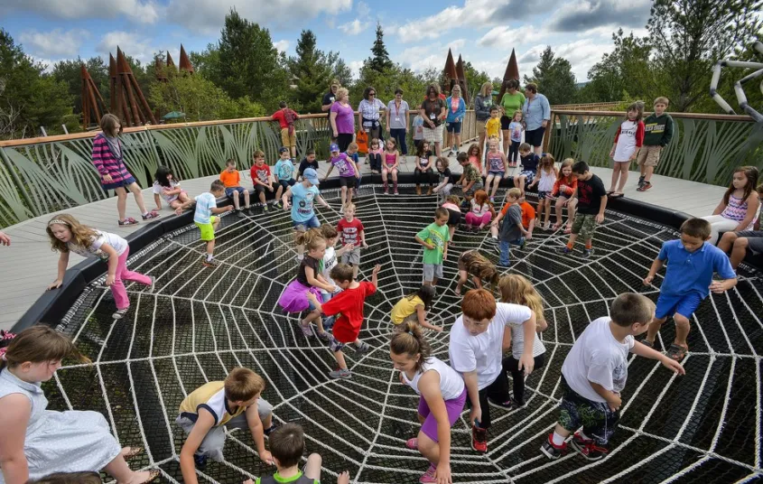 People crossing a suspended metal walkway between large wooden structures at The Wild Center in Tupper Lake NY 