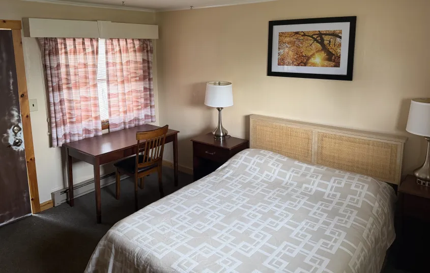 A king bed in a room with a desk and beige walls at Tupper Lake Motel.