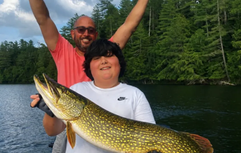 A kid holds a large pike while a man throws up his hands in victory behind.