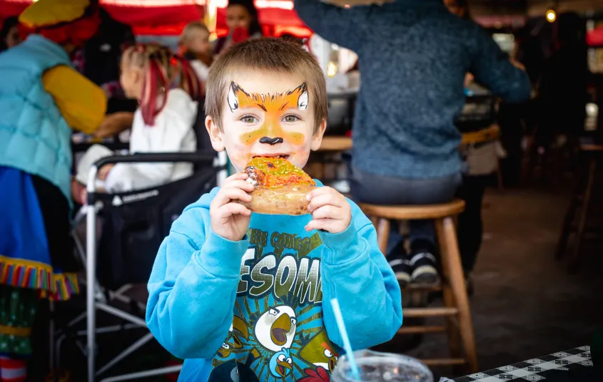 A young boy with his face painted like a fox enjoys a slice of pizza .
