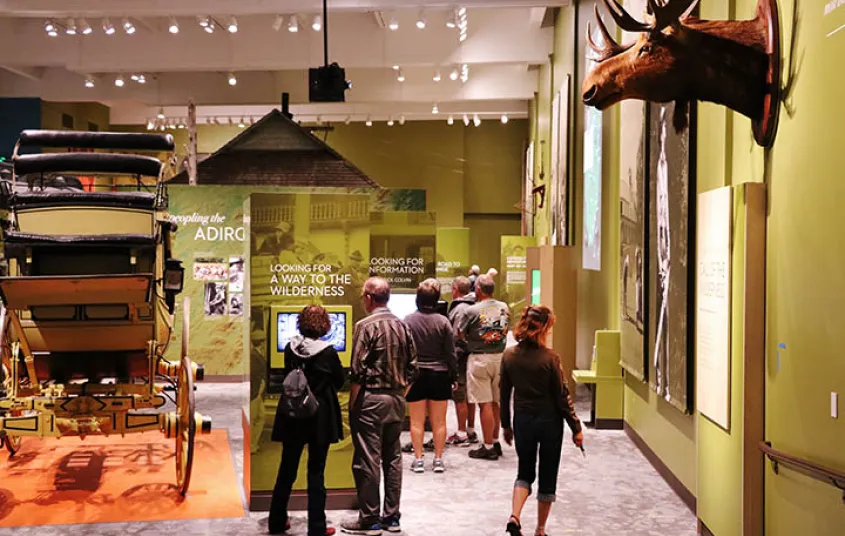 A large room with green walls and high ceilings where people are viewing the plaques under various displays like a carriage and a moose.