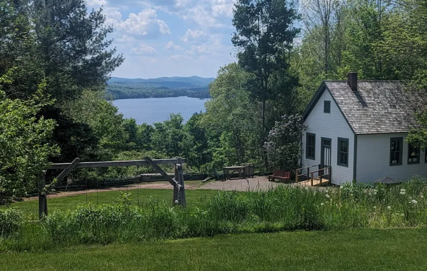 Blue Mountain Lake under billowing white clouds across a lush green lawn in front of a quaint white cabin.