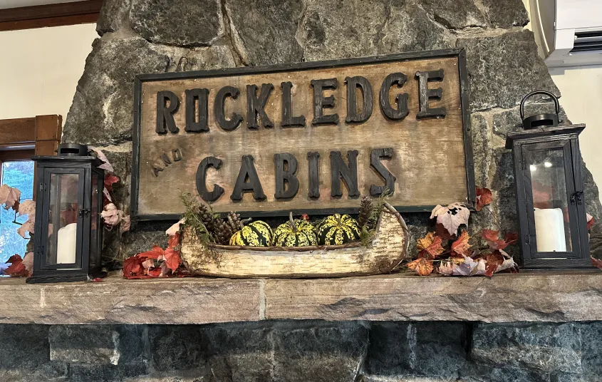 A carved sign reading 'Rockledge and Cabins' over a stone mantle with birch bark boat filled with festive squash and candles.