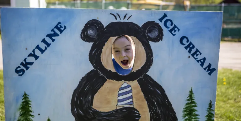 A child smiles through the hole of the cut out with a bear holding an ice cream