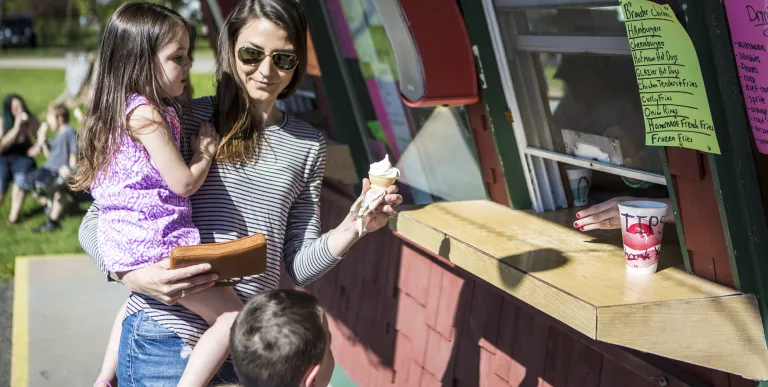 A mom holds her kid in one hand and an ice cream cone in the other.