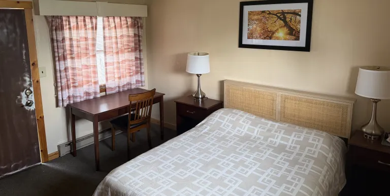 A king bed in a room with a desk and beige walls at Tupper Lake Motel.