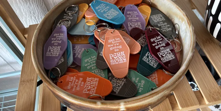 A takeaway paper cup with custom Perk and Pine cardboard sleeve on a wooden window sill next to a plant potted in a disco ball