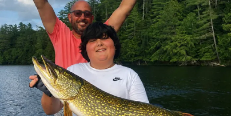 A kid holds a large pike while a man throws up his hands in victory behind.