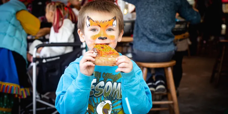 A young boy with his face painted like a fox enjoys a slice of pizza .