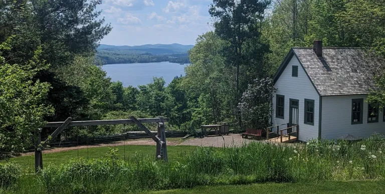 Blue Mountain Lake under billowing white clouds across a lush green lawn in front of a quaint white cabin.