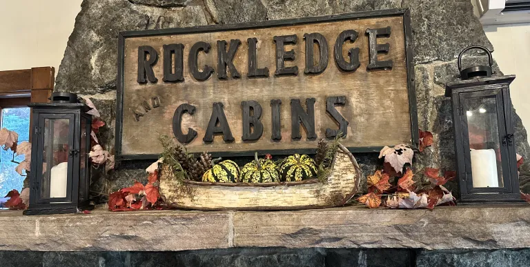 A carved sign reading 'Rockledge and Cabins' over a stone mantle with birch bark boat filled with festive squash and candles.